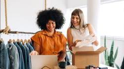 Two women happily preparing clothing items for shipment in a small business setting.